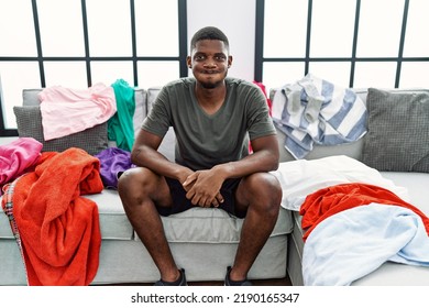 Young African American Man Sitting On The Sofa With Dirty Laundry Clothes Puffing Cheeks With Funny Face. Mouth Inflated With Air, Crazy Expression. 