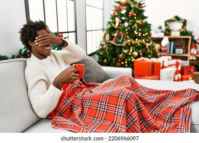 Young African American Man Sitting On The Sofa Drinking Coffee By Christmas Tree Smiling And Laughing With Hand On Face Covering Eyes For Surprise. Blind Concept. 