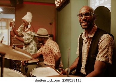 Young African American man sitting at drum kit and smiling at camera while two musicians getting ready for performance in background - Powered by Shutterstock