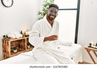 Young African American Man Smiling Happy With Facial Mask Drinking Champagne At Beauty Center.