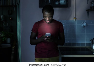 Young African American Man Standing In His Kitchen Using Smartphone, Browsing Online Or Exchanging Messages With Friend With Happy Confident Smile At Night