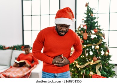 Young African American Man Standing By Christmas Tree With Hand On Stomach Because Indigestion, Painful Illness Feeling Unwell. Ache Concept. 