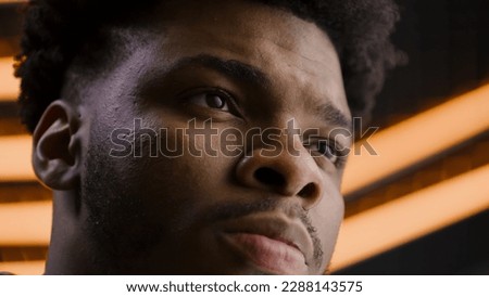 Young African American man straight and serious face with confident deep look. Focused worker looks away, blinks eyes, poses on camera. LED lamps on background in studio or office. Close up portrait.