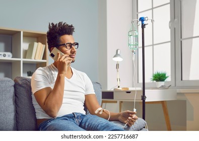 Young African American man talking with his doctor on his mobile phone while sitting on the sofa at home and receiving medication through an intravenous drip - Powered by Shutterstock