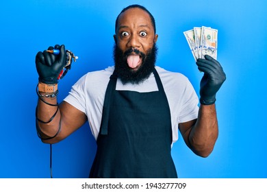 Young African American Man Tattoo Artist Wearing Professional Uniform And Gloves Holding Tattooer Machine And Usa Dollars Banknotes Sticking Tongue Out Happy With Funny Expression. 