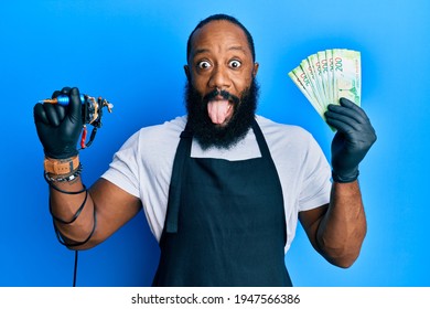 Young African American Man Tattoo Artist Wearing Professional Uniform And Gloves Holding Tattooer Machine And Russian Ruble Banknotes Sticking Tongue Out Happy With Funny Expression. 