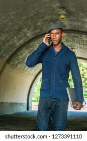 Young African American Man Traveling In New York City, Wearing Blue Shawl Collar Cardigan Sweater, Gray Patterned Flat Cap, Holding Laptop Computer, Talking On Cell Phone, Walking Under Street Bridge.