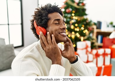 Young African American Man Using Headphones Sitting By Christmas Tree At Home.