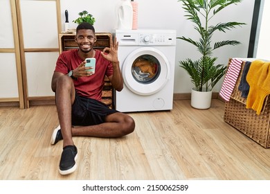 Young african american man using smartphone waiting for washing machine smiling positive doing ok sign with hand and fingers. successful expression. 
