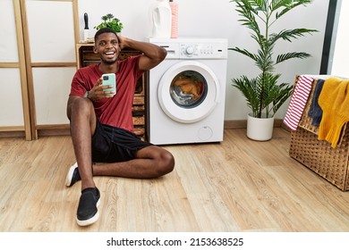 Young African American Man Using Smartphone Waiting For Washing Machine Smiling Confident Touching Hair With Hand Up Gesture, Posing Attractive And Fashionable 