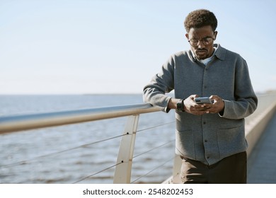 Young African American man using smartphone while standing near a waterfront railing on a bright day. Focused expression on man's face while browsing on his device - Powered by Shutterstock