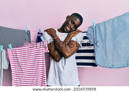 Young african american man washing clothes at clothesline hugging oneself happy and positive, smiling confident. self love and self care 
