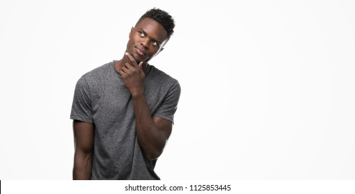 Young african american man wearing grey t-shirt with hand on chin thinking about question, pensive expression. Smiling with thoughtful face. Doubt concept.