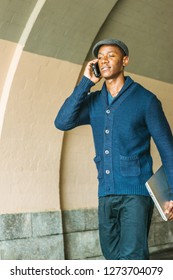 Young African American Man Wearing  Blue Shawl Collar Cardigan Sweater, Black Pants, Flat Cap, Holding Laptop Computer, Talking On Cell Phone, Walking Through Under Street Bridge In New York City.
