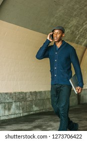 Young African American Man Wearing  Blue Shawl Collar Cardigan Sweater, Black Pants, Flat Cap, Holding Laptop Computer, Talking On Cell Phone, Walking Through Under Street Bridge In New York City.
