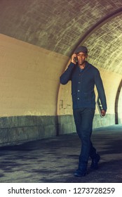 Young African American Man Wearing  Blue Shawl Collar Cardigan Sweater, Black Pants, Sneakers, Flat Cap, Holding Laptop Computer, Talking On Cell Phone, Walking Through Under Street Bridge In New York