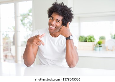 Young African American Man Wearing Casual White T-shirt Sitting At Home Smiling Doing Talking On The Telephone Gesture And Pointing To You. Call Me.