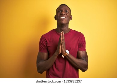 Young African American Man Wearing Red T-shirt Standing Over Isolated Yellow Background Begging And Praying With Hands Together With Hope Expression On Face Very Emotional And Worried. Asking