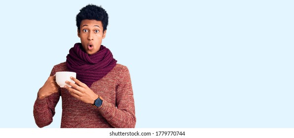Young african american man wearing winter scarf and drinking a cup of hot coffee scared and amazed with open mouth for surprise, disbelief face 