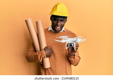 Young african american man wearing safety helmet holding blueprints and drone smiling and laughing hard out loud because funny crazy joke. 