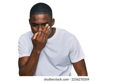 Young African American Man Wearing Casual White T Shirt Smelling Something Stinky And Disgusting, Intolerable Smell, Holding Breath With Fingers On Nose. Bad Smell 