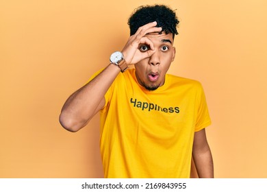 Young african american man wearing t shirt with happiness word message doing ok gesture shocked with surprised face, eye looking through fingers. unbelieving expression. 