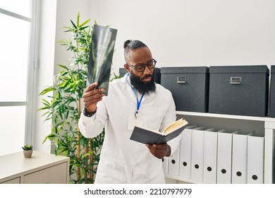 Young African American Man Wearing Doctor Uniform Reading Book Looking Xray At Clinic