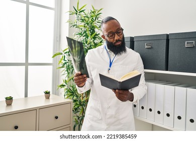 Young African American Man Wearing Doctor Uniform Reading Book Looking Xray At Clinic