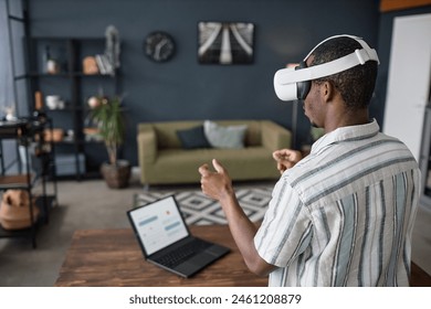 Young African American man wearing VR headset standing in living room surfing Internet on laptop, copy space - Powered by Shutterstock