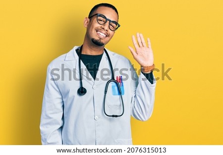 Young african american man wearing doctor uniform and stethoscope waiving saying hello happy and smiling, friendly welcome gesture 