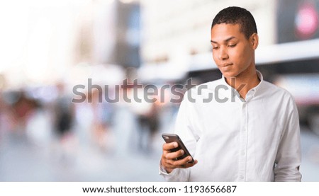 Young african american man with white shirt sending a message or email with the mobile in the middle of the city