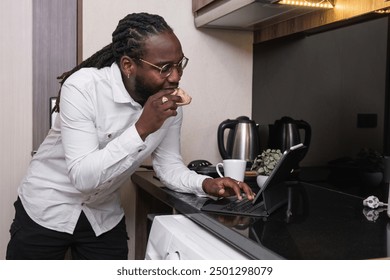 Young African American Man Working Online in Modern Kitchen, Using Laptop, Eating Breakfast, Remote Work, Home Office, Casual Attire, Technology, Lifestyle - Powered by Shutterstock