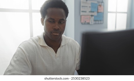 Young african american man working indoors in an office setting, focusing on his computer with a window and bulletin board in the background - Powered by Shutterstock