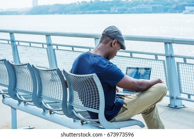 Young African American Man working on laptop computer outdoor in New York, wearing blue T shirt, flat cap, crossing legs, sitting on metal chair by Hudson River, looking down, reading. Back View. 
