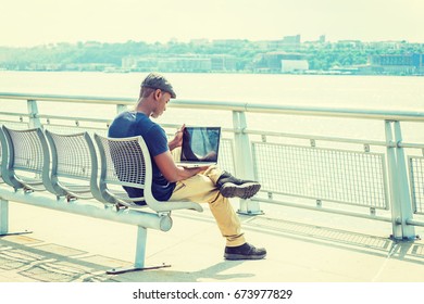 Young African American Man working on laptop computer outdoor in New York, wearing blue T shirt, flat cap, crossing legs, sitting on metal chair by Hudson River, looking down, reading. Back View. 
