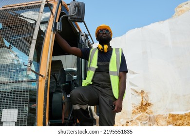 Young African American man in workwear and hardhat standing in excavator or other construction machine during work