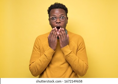 Young African American Man In Yellow Sweater In Shock, Keeping Mouth Wide Open And Hands Near Mouth, Feeling Stressed After Having Forgotten To Pay Bills In Time. Studio Shot