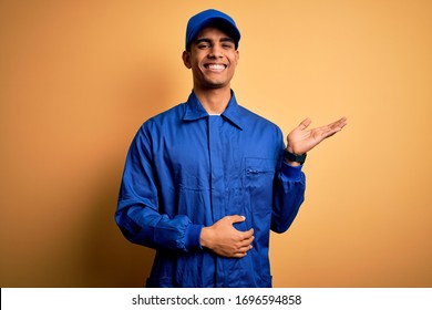 Young african american mechanic man wearing blue uniform and cap over yellow background smiling cheerful presenting and pointing with palm of hand looking at the camera.