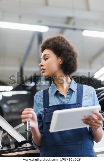 young african american mechanic in\
overalls holding car key and digital tablet in\
garage