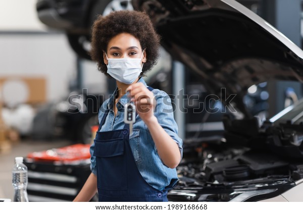 young african american mechanic in\
protective mask holding blurred car key in hand in\
garage