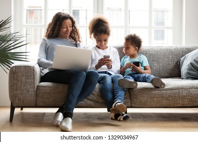 Young African American Mom And Small Kids Sit On Couch Using Devices, Busy With Laptop And Smartphones, Black Modern Family Addicted To Gadgets, Little Children Play On Cellphone. Technology Concept