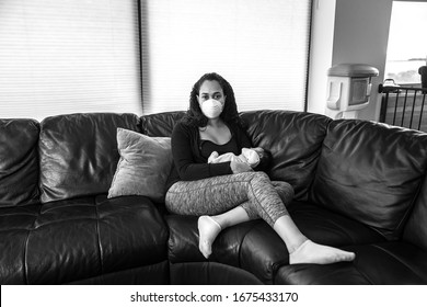 A Young African American Mother Sits On A Black Leather Sofa With Legs Crossed Holding Her Newborn Daughter Wearing A Dust Face Mask In Hopes Of Preventing Getting Sick From Caronavirus Or COVID-19.