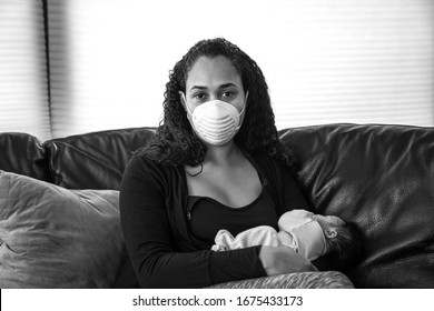 A young African American mother sits on a black leather sofa holding her newborn daughter to her chest wearing a dust mask on her face in hopes of preventing sickness from caronavirus or COVID-19.