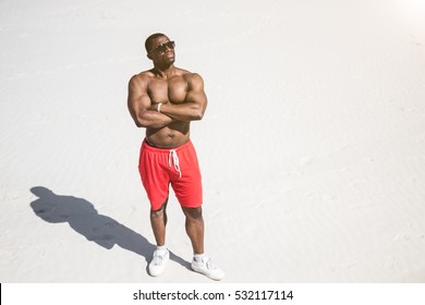 Young African American Muscle Man In Red Shorts And Sunglasses Standing In The Sand On The Beach And Looking Up