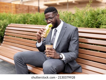 Young African American Office Worker Having Lunch Outdoors, Drinking Coffee And Eating Sandwich While Sitting On Bench Near Office Building, Copy Space