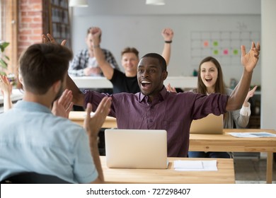 Young African American Office Worker Throws Hands In Air Celebrating Achievement At Work. Coworkers Around Cheering And Clapping Hands. Rewarding Outcome, Received Promotion, Achieved Success Concept.