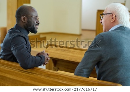 Young African American pastor consulting senior male parishioner after church service while both sitting on bench in front of one another