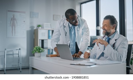 Young African American Physician Is Talking To A Senior Doctor In A Medical Health Clinic. Colleagues Discuss Work On Laptop Computer In Hospital Office. Assistant Asks Medical Advice From A Doctor.