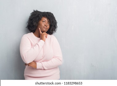 Young african american plus size woman over grey grunge wall wearing winter sweater with hand on chin thinking about question, pensive expression. Smiling with thoughtful face. Doubt concept. - Powered by Shutterstock