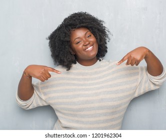 Young african american plus size woman over grey grunge wall wearing a sweater looking confident with smile on face, pointing oneself with fingers proud and happy.
